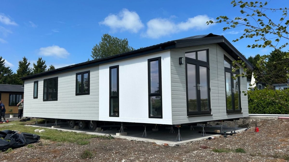 A modern single-storey modular home with light grey cladding and black-framed windows, elevated on supports during installation, set against a clear blue sky with surrounding greenery.