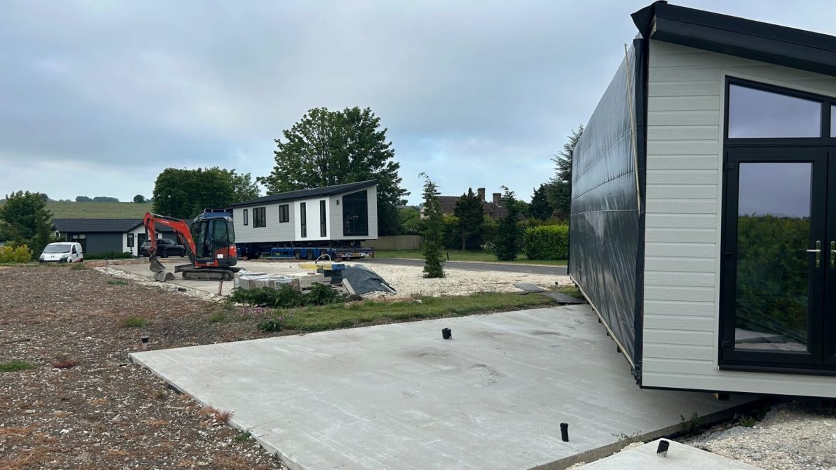 A modular home installation site with one lodge section on a transport trailer in the background, a second section positioned in the foreground, and a small excavator and construction materials on site, surrounded by greenery under an overcast sky.