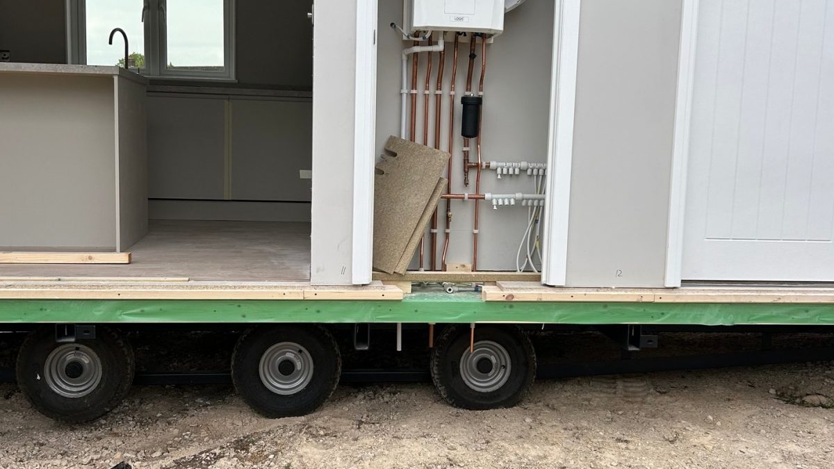Close-up view of a modular home section during installation, showing exposed undercarriage wheels, kitchen interior on the left, and boiler with copper pipework in a utility cupboard on the right