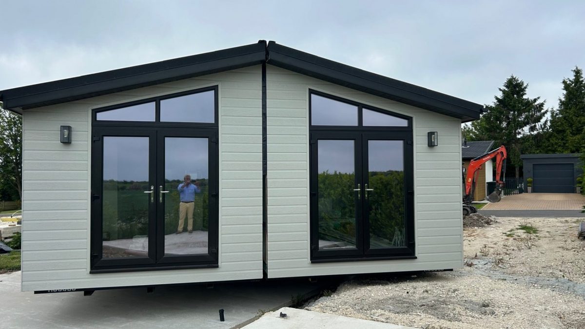 Front view of a modular home during installation, showing two connected sections with light grey cladding, black-framed double doors, and large windows, with construction equipment and a garage visible in the background.