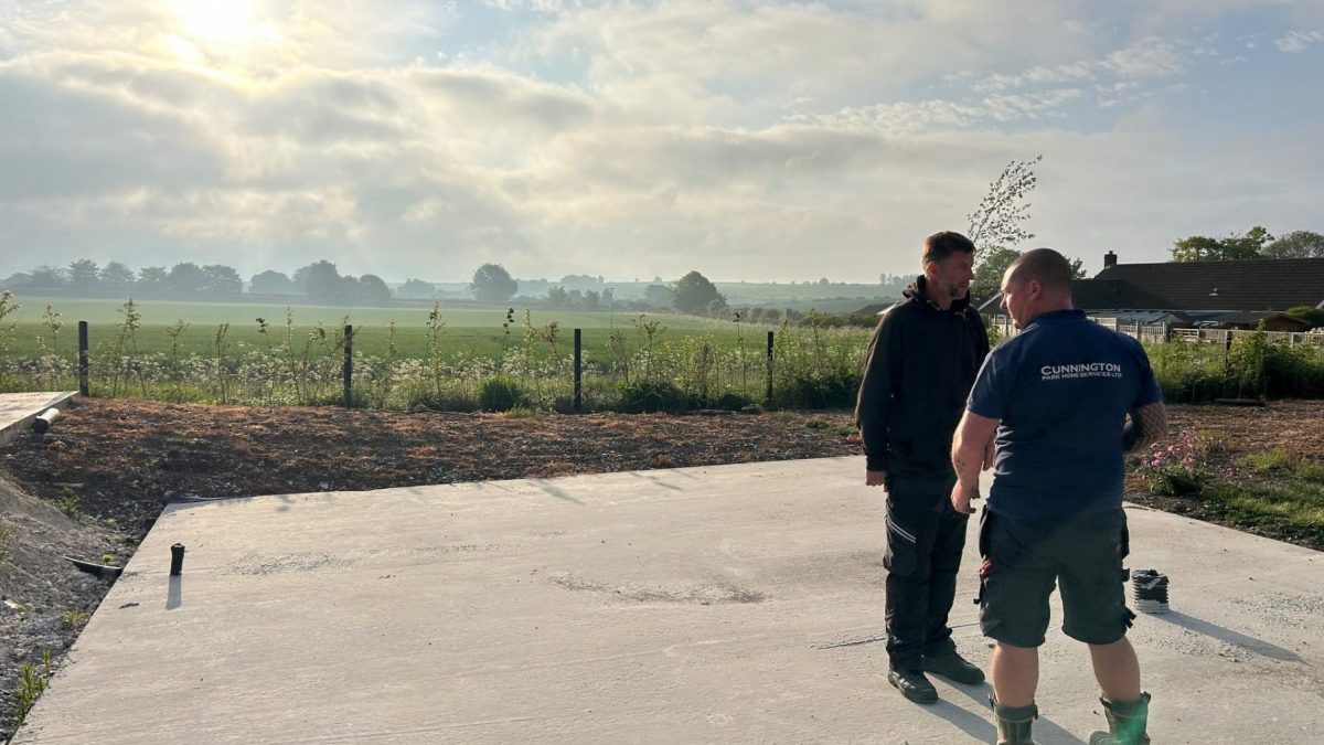 wo workers standing on a newly laid concrete base at a construction site in the early morning, with open countryside and a hazy sunrise in the background.