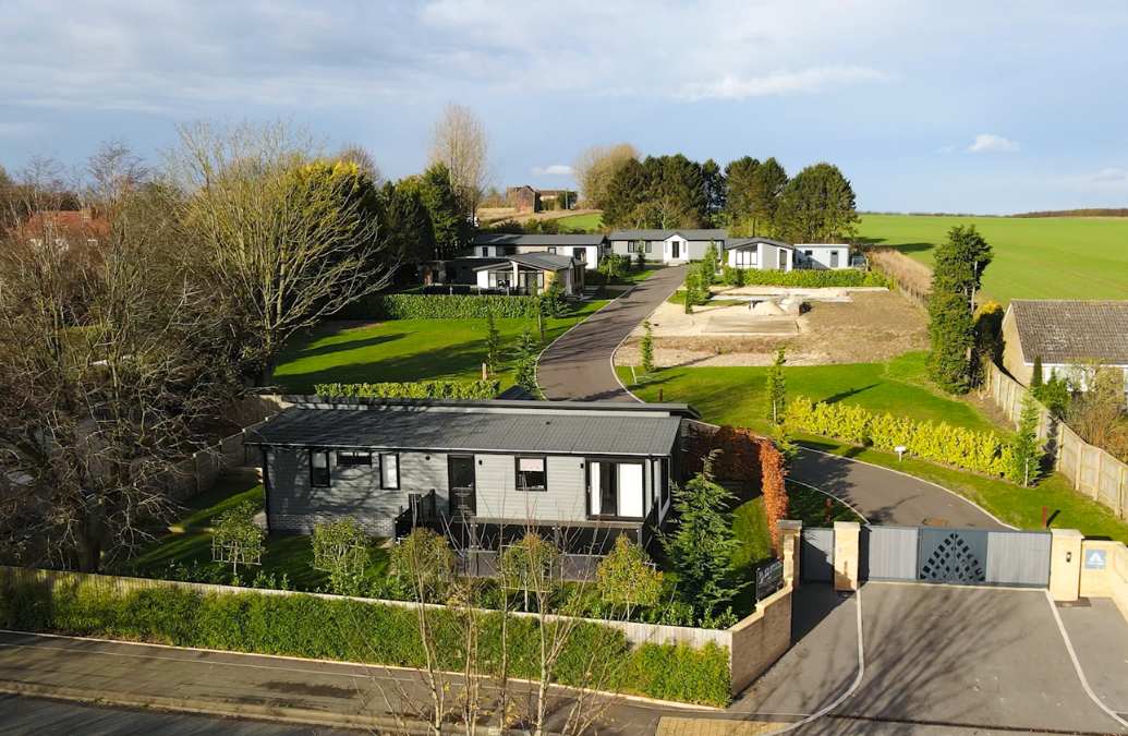 A gated residential development with landscaped gardens, modern single-storey homes, and a winding private driveway, surrounded by green fields and trees under a partly cloudy sky